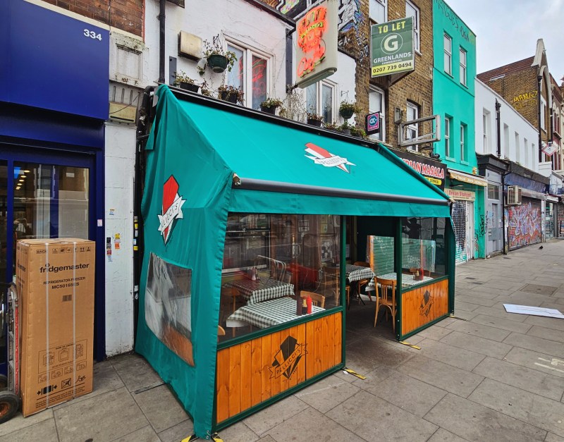 Exterior view of a restaurant with a green awning and wooden paneling, featuring glass walls and outdoor seating.