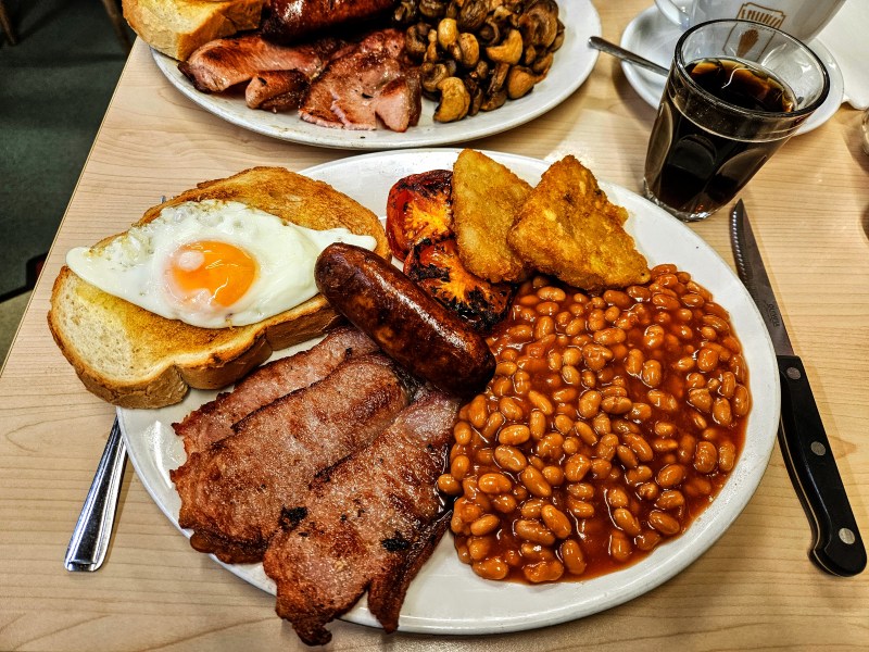 A full English breakfast served on a white plate, featuring crispy bacon, a fried egg, sausage, baked beans, hash browns, grilled tomatoes, and toast, with a glass of Coke on the side.