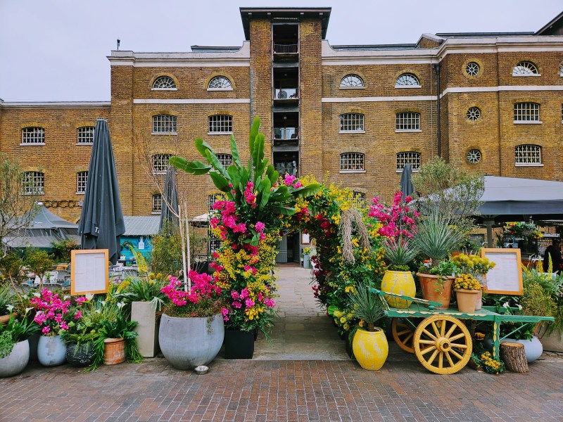 Entrance to a vibrant outdoor area featuring colorful flowers and plants, with a brick building in the background.