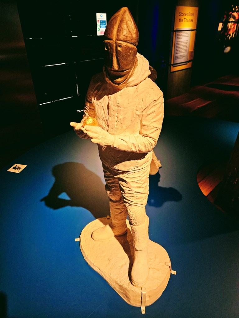 A clay statue of a historical figure, wearing a helmet and holding an object, displayed in a museum setting with dim lighting and informative signage in the background.