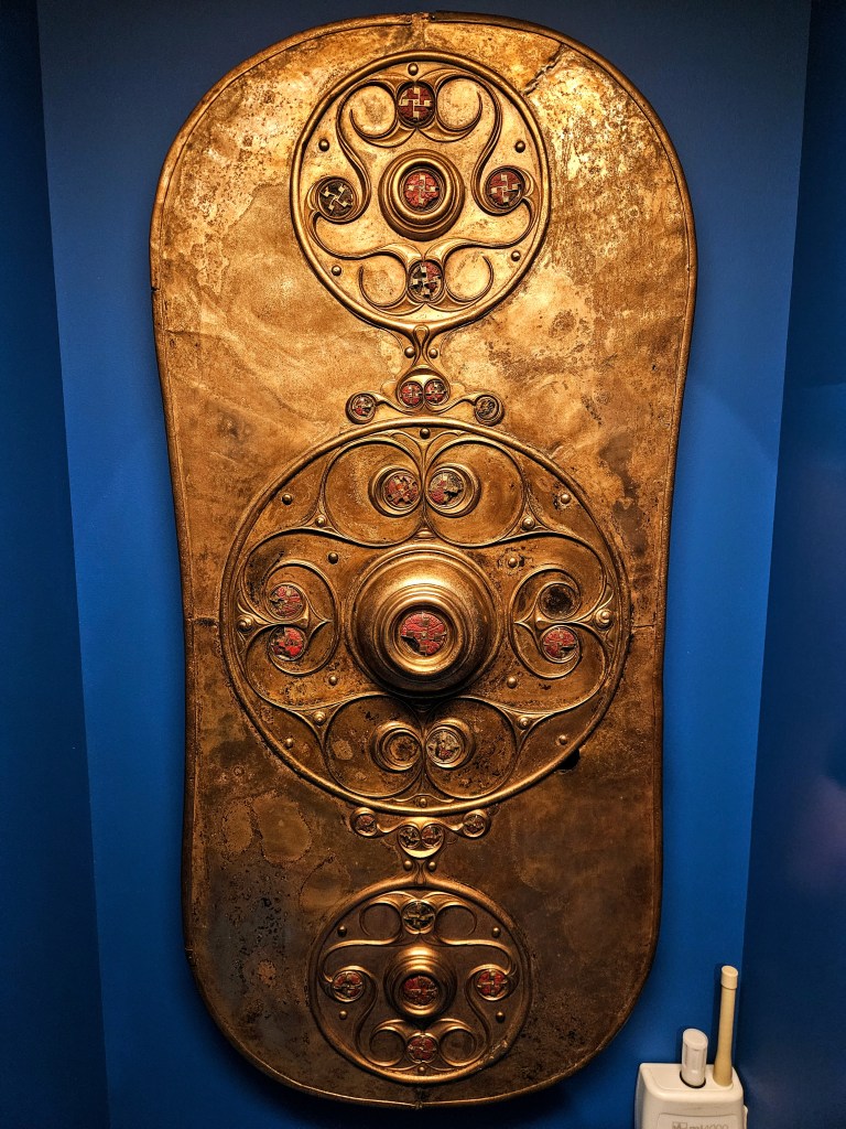 A decorative golden plate with intricate circular patterns and colored insets, displayed against a blue wall.