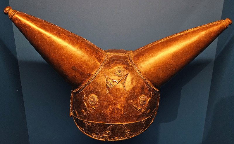 A decorative historical helmet with two prominent, cone-shaped horns, displayed against a blue background.