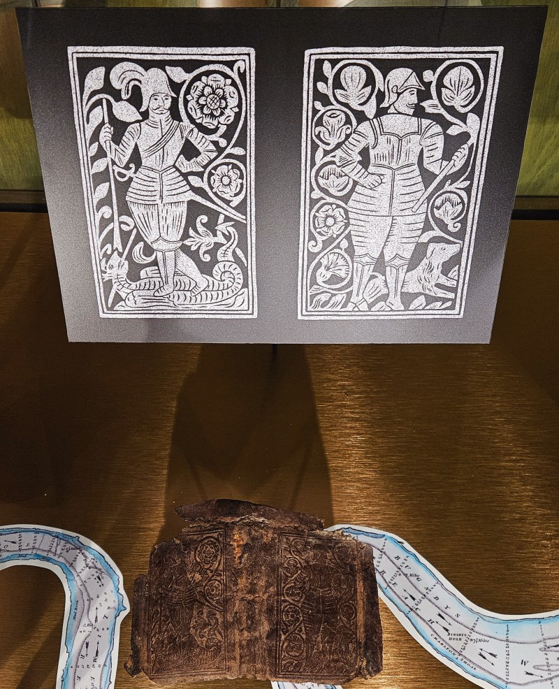 Two illustrated panels depicting historical figures surrounded by floral designs, displayed alongside a worn leather item and a map fragment.