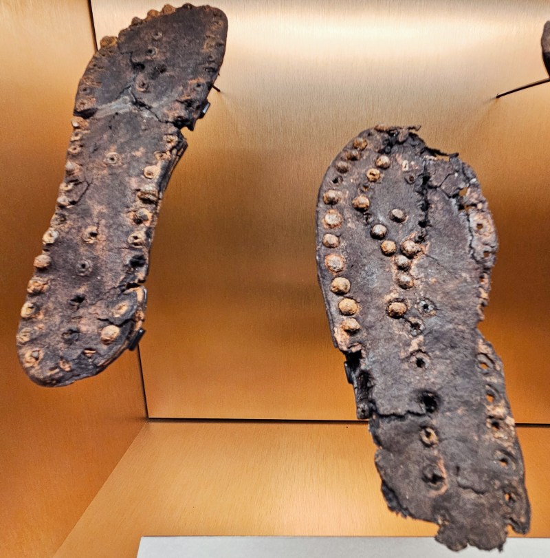 A pair of ancient sandals displayed in a museum exhibit, featuring a worn-out design with circular studs along the edges.
