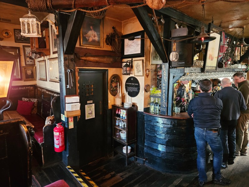 Interior of a cozy pub, featuring wooden beams, framed artwork on the walls, and patrons at the bar. A small seating area with cushions can be seen in the corner.