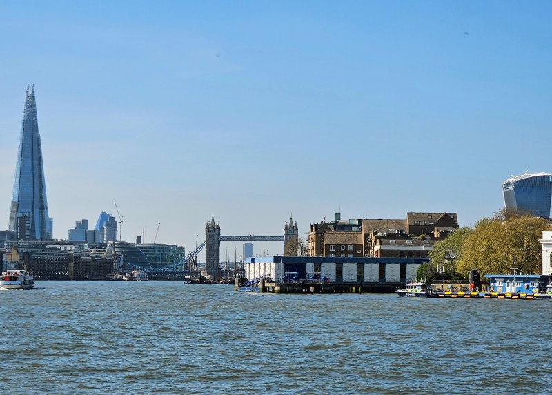 A view of the River Thames with the Tower Bridge in the distance, featuring modern and historical buildings along the waterfront on a sunny day.