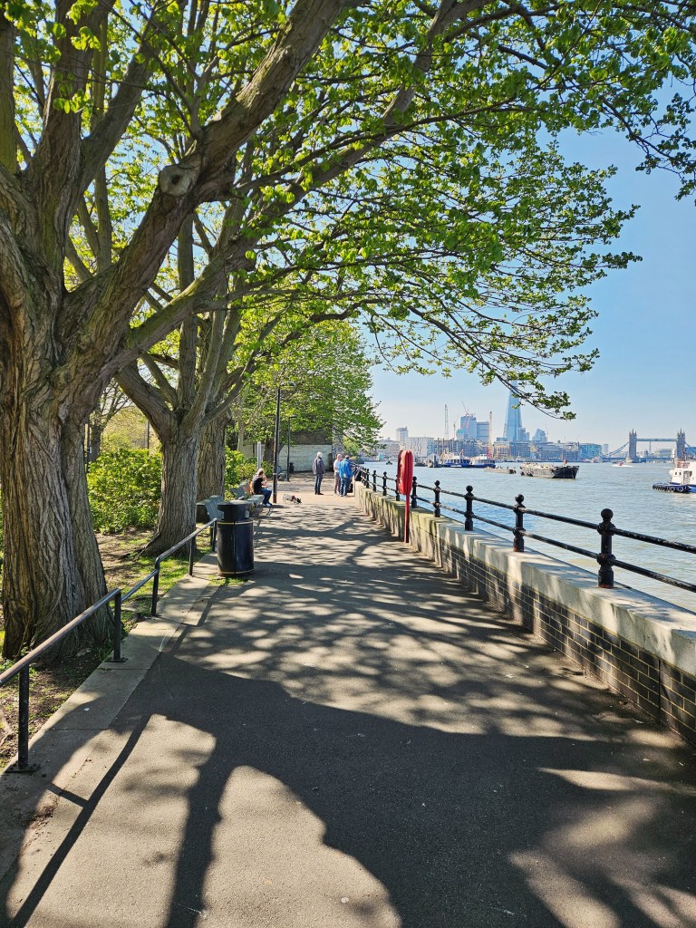 A scenic riverside path lined with green trees and a black railing, showing a sunny day with people walking and enjoying the view of the water.