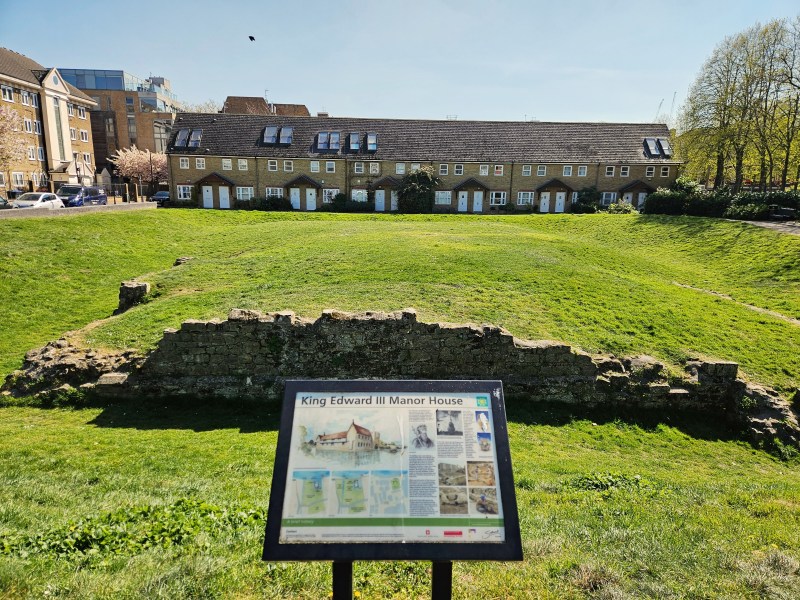View of the ruins of King Edward III Manor House with a nearby informational sign, set against a backdrop of modern buildings and green grass.