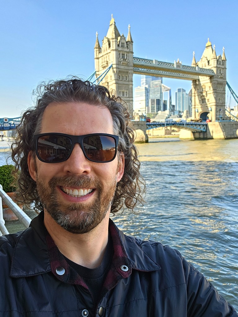 A person smiling in front of Tower Bridge on a sunny day, wearing sunglasses with the River Thames in the background.