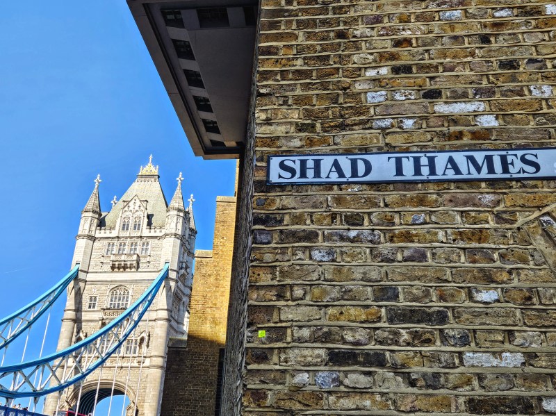 A view of Tower Bridge in London peeking over a brick wall with a street sign that reads 'SHAD THAMES'.