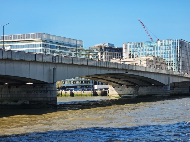A view of London Bridge spanning the River Thames, with modern buildings in the background and a clear blue sky.