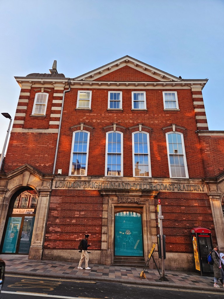 A historic brick building with large windows, featuring the engraved words 'LONDON BRIGHTON AND SOUTH COAST RAILWAY' on the facade. A person walks by on the sidewalk, and a red telephone booth is visible nearby.