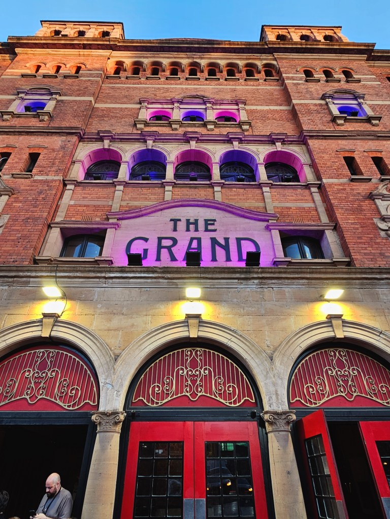 The exterior of a theater building named 'The Grand', featuring red doors and decorative arches, illuminated with purple lighting against a clear blue sky.